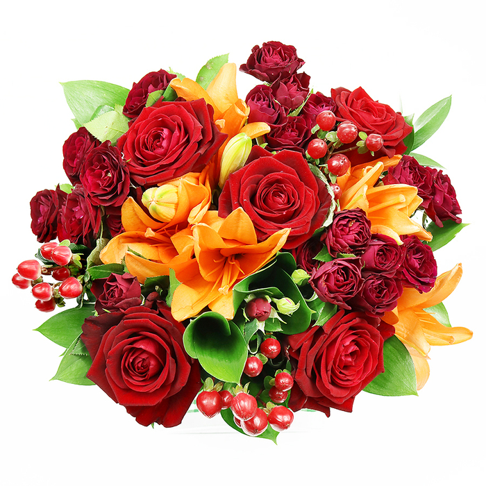 Captured from above on a spotless white field, this lavish, circular bouquet reads like a jewel box of autumnal romance. Large, fully blossomed deep red roses anchor the design, each bloom showing velvety, layered petals that open toward their darker, textured centers-an image of classic devotion. Interspersed among them are vivid orange Asiatic lilies whose trumpet-formed petals and exposed stamens introduce architectural interest and bright, optimistic warmth; a few lilies remain partially closed or in tight bud, suggesting freshness and staged unfolding. Nestled in the spaces between these principal flowers are clusters of small, dark red spray roses, their compact faces contributing a plush, opulent depth that edges the colour scheme toward wine tones. Scattered throughout are glossy hypericum berries-ruby spheres that reflect light and add a playful, tactile element to the composition. Lush green foliage, likely ruscus or salal, frames the blooms with broad, shiny leaves that both contrast and harmonise with the red and orange hues. The lighting is gentle and even, chosen to reveal petal texture and berry sheen without dramatic shadow, offering a clear sense of volume and craftsmanship. This bouquet imparts sensory richness-visual warmth, the soft density of rose petals, a suggestion of lily perfume-and is ideal for marking special moments such as anniversaries, intimate celebrations, or to bring seasonal colour to a home or table a short walk from Elmers End station or the nearby green spaces. The overall effect is one of considered abundance and heartfelt style.