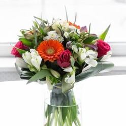 A breathtaking floral showcase reminiscent of Elmers End's most inviting garden corners is artfully displayed in this arrangement, with soft daylight casting a serene glow. The bouquet's heart is formed by creamy-beige and vibrant orange gerberas, their round, joyful faces echoing the gentle optimism of a sunny morning in nearby Beckenham. Tightly wrapped fuchsia roses nestle thoughtfully among them, their deep colour lending richness and depth, while clusters of pristine white alstroemeria and pepperings of soft pink and white filler blooms add a lyrical lightness, much like spring blossoms seen in South Norwood Country Park. The lushness of the design is grounded by wide, glossy aspidistra leaves, artfully shaped into a protective cradle around the base, offering both structure and a lush, tranquil feel that channels the calm of a local park's green spaces. Slender, pointed greenery and small sprigs create gentle separation between blooms, further enhancing the bouquet's fullness and vitality. Set within a slender glass vase that reveals the vibrant, freshly cut stems, this composition is both classical and modern-perfectly tailored for gatherings, hospitality, or welcoming someone special to a new chapter in the Elmers End area. The arrangement embodies a sense of harmony and hope, crafted by local floral experts whose connection to community resonates in every stem and petal.